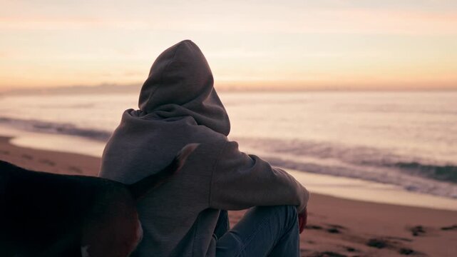 Solitary man in a hoodie sitting with his loyal dog on a sandy beach at sunset, peacefully watching the gentle waves and enjoying the serene coastal landscape together in a quiet moment
