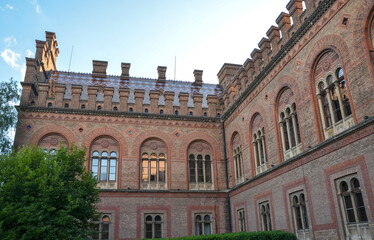 Fototapeta premium Stunning red brick architecture of Chernivtsi National University features ornate masonry and arched windows. A decorative tile roof and a central domed tower stand out against a clear blue sky