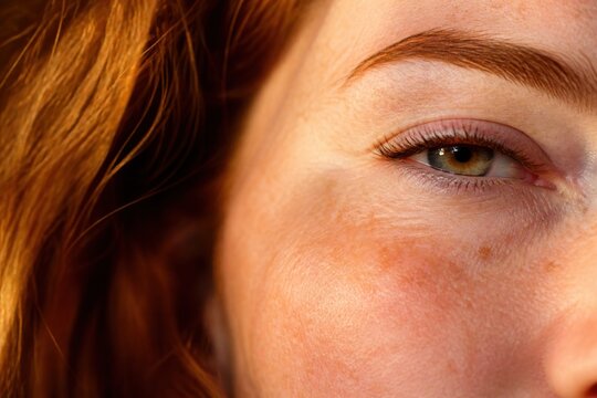 Close up of a red-haired woman's eye with freckles on her cheek. The warm golden light highlights the natural skin texture and ginger hair. Ideal for skincare or beauty advertising.