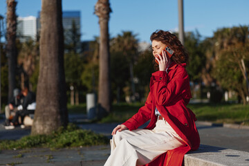 Obraz premium Young woman with curly hair wearing a red jacket talks on a smartphone while sitting on a bench in a sunny urban park with palm trees in the background.