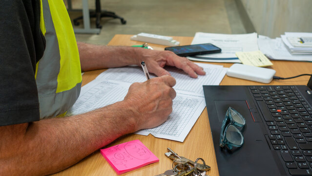 Construction manager in high visibility vest at his desk filling out work and safety report cards. Engineer at his work station with a laptop signing off on his work sheets.