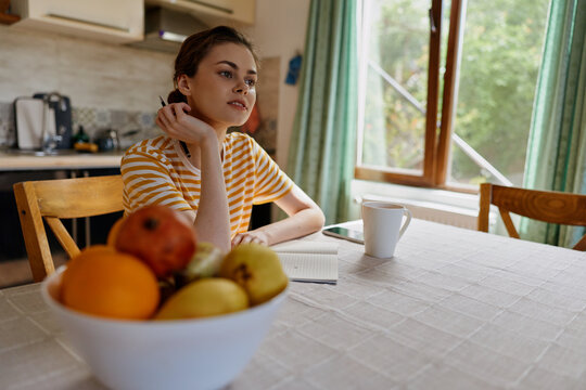 A young woman in a striped yellow top sits at a sunlit kitchen table with a bowl of fruit beside her, gazing thoughtfully out the window as soft light fills the room.