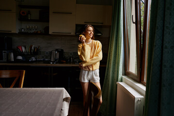 Obraz premium A woman in a cozy kitchen by a sunlit window, wearing a yellow sweater and shorts, smiles while holding an orange. A warm home lifestyle moment.