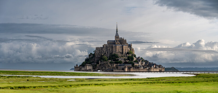 Road to Mont-Saint-Michel during high tide with rising water, tidal phenomenon on Atlantic's ocean coast, view on UNESCO heritage site and iconic Normandy landmark and green pastures