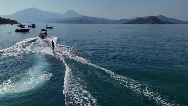 A wakeboarder rides behind a speedboat on calm blue sea, with mountain islands and white boat wake in a sunny coastal landscape, ideal for travel, adventure and water sport themes.