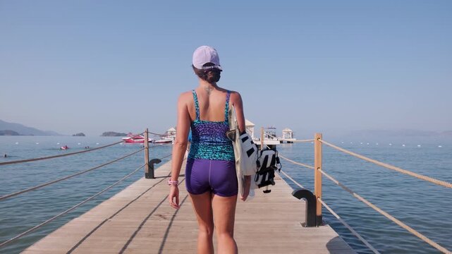Rear view of a woman carrying wakeboard gear on a wooden pier by calm blue sea, with boats and islands ahead, creating a sunny summer mood for travel, leisure and water sport preparation themes.