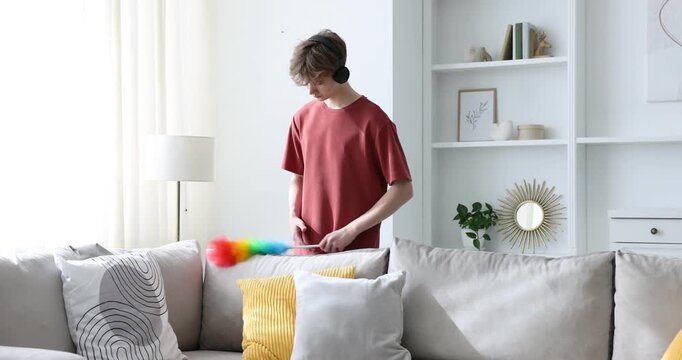 Housework. Teenage boy in headphones removing dust from pillows with static duster at home