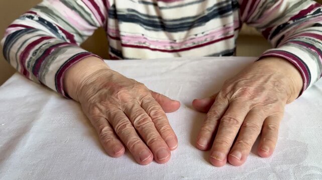 Close view of an elderly woman's hands and forearms resting palm-down on a white textured tablecloth. Striped long-sleeve shirt sleeves frame visible veins
