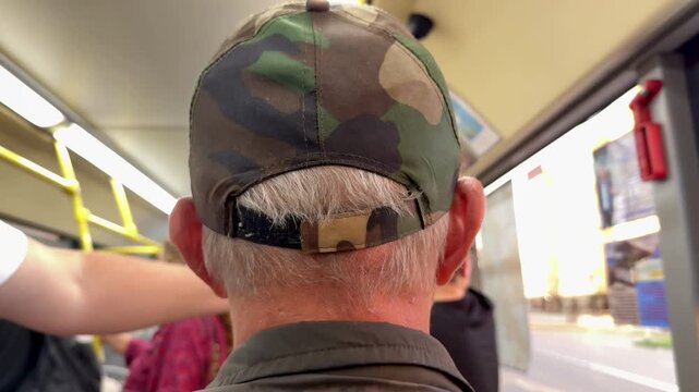 Rear view white man on bus seat wearing camouflage cap and gray jacket close-up of head and shoulders crowded interior daytime