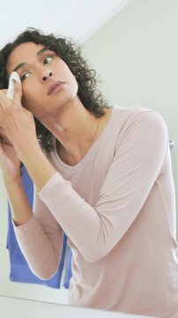 Vertical video: Grabbing white wipe, non-binary person removing makeup at bathroom mirror, pink top