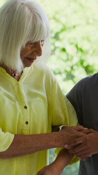 Vertical video: Reaching senior seeking comfort while caregiver holding hand by home window, scrubs