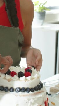 Vertical video: Squeezing piping bag baker in green apron finishing 2tier cake with berries at home