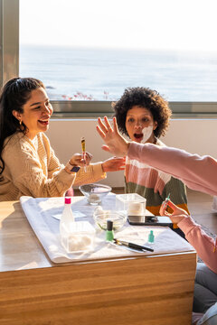 Diverse family with child applying cream and painting nails on towel at table by sea window