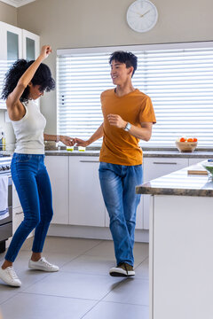 Diverse couple dancing by island bowl of oranges, blinds filtering daylight and inspiring movement