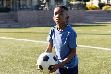 Naklejka premium African American child holding black-white soccer ball on turf in blue tee shorts looking right