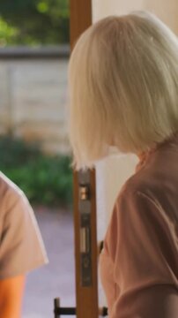 Vertical video: Seeing visitor appearing senior woman in pink top opening door at home touching arm