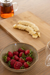 Ceramic bowl holding strawberries is sitting on wooden table near glass teapot and banana slices © wavebreak3
