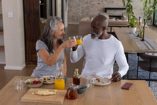 Diverse mature couple raising clear glasses of orange juice at home dining table with glass pitcher