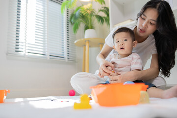 Fototapeta premium Warm sunlight bathes a cozy playroom scene. Mother gently holds her infant, eyes full of affection. Soft toys and bright containers dot the floor nearby. Natural light filters through window blinds 