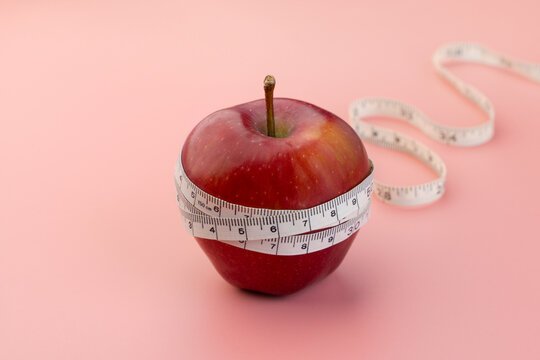 A red apple wrapped in a white measuring tape on a pink background. Weight loss, dieting, nutrition, and healthy lifestyle goals concept. Copy space for text.