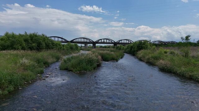 River flows beneath a long metal bridge. Grassy banks frame the water's path. Sky shows clouds drifting above. Bridge arches stretch across the scene. Water rushes gently over stones