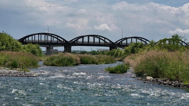 Rapid river flows beneath a long, arched bridge. Green grass and reeds line the banks. Sky holds soft clouds above. Bridge spans the rushing water with sturdy arches
