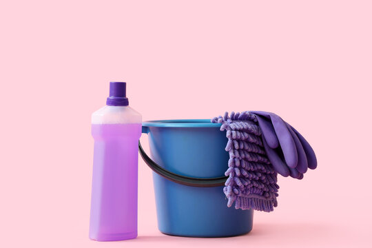 Household cleaning supplies with purple detergent bottle blue bucket microfiber mop and rubber gloves on pink background