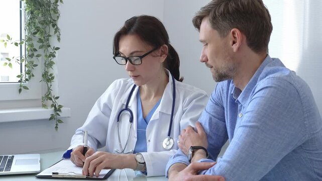 Female doctor taking notes on clipboard while listening attentively to patient during medical consultation in hospital office, promoting communication and understanding.