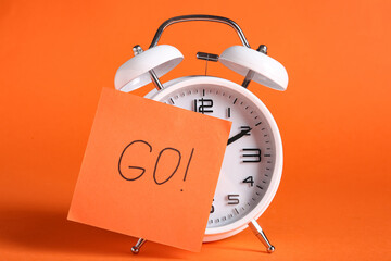 Alarm clock with word GO on sticky note against orange background. Time management concept