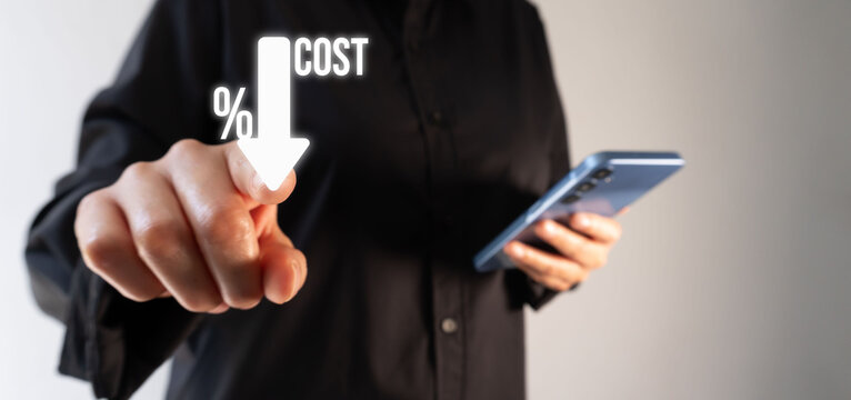 Person points to digital graphic showing falling cost percentage. Hand gestures toward arrow symbolizing reduction. Holds smartphone while focusing on cost-saving concept.