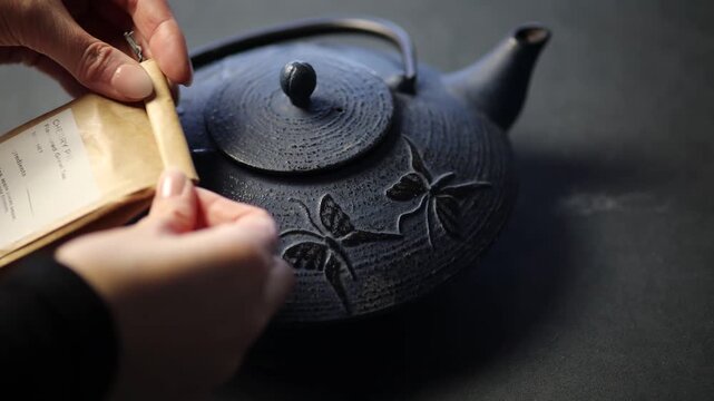 hands gently opening a paper tea bag followed by opening a cast iron teapot. The scene emphasises calm, mindful tea preparation with soft, deliberate movements in a traditional tea setting.