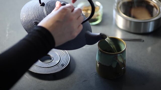 Hands pouring freshly brewed tea into a ceramic cup in a calm kitchen setting. The scene highlights a simple tea preparation moment with warm liquid flow, natural lighting.