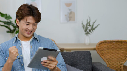 Excited Young Asian Man Celebrating Success While Using Digital Tablet on Sofa © blueringmedia