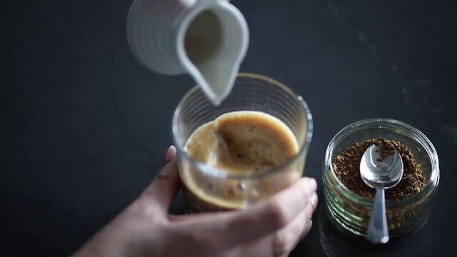 Hands pouring milk from a jug into a glass cup containing freshly frothed instant coffee mixture.