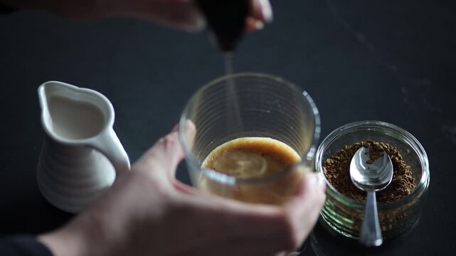 hands using an electric frother stick to blend and froth a mixture of instant coffee, honey, and hot water in a glass cup. The liquid transforms into a smooth foam, highlighting a homemade coffee.