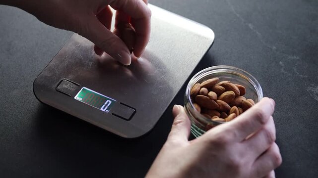 Close-up video showing hands counting almonds and placing them one by one onto electronic kitchen scales in a home kitchen setting. The scene highlights careful portion control.