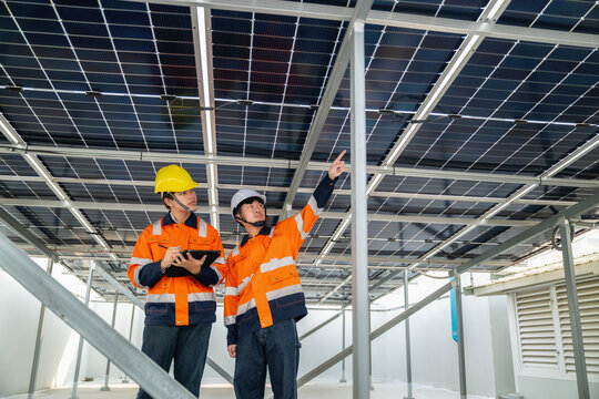 Young technician and Engineer inspect solar panels on steel framework on building rooftop. One points upward, the other holds a tablet. They wear bright orange safety vests and hard hats.