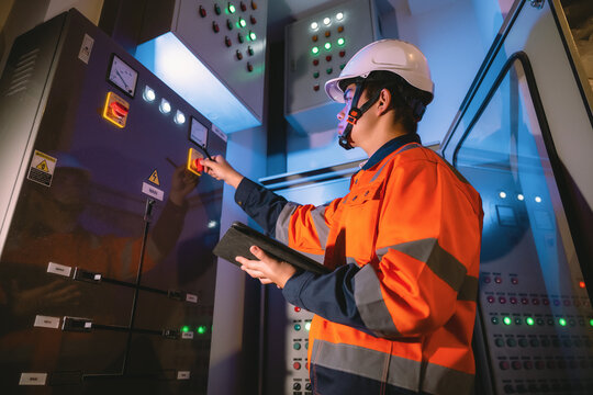 Professional engineer in high visibility jacket controlling electricity cabinet, holding tablet operating main power supply switch, operating breaker toggle on industrial electrical board.