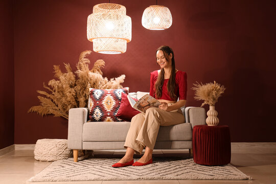 Smiling woman reading magazine on sofa in cozy living room with bohemian decor and warm lighting