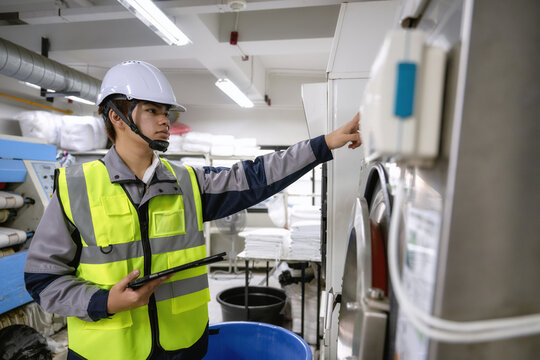 Maintenance technician holding tablet operating industrial laundry machine control, checking large washing machine factory subsystem, adjusting settings on large industrial washing appliance.