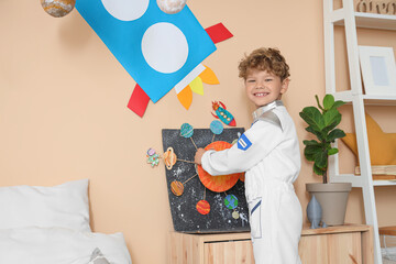 Smiling young boy in astronaut costume playing with solar system model and rocket decorations in bedroom, the bedroom features colorful rocket decorations on the wall © Pixel-Shot