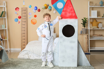 Smiling young boy in astronaut costume posing with cardboard rocket ship in space-themed bedroom, the room features a growth chart decorated with planets and rockets, emphasizing childhood © Pixel-Shot