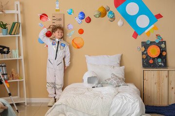 Happy young boy in astronaut costume measuring height in space-themed bedroom with rocket and planet decorations, the room features rocket and planet decorations © Pixel-Shot