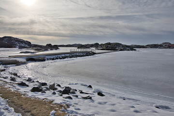 Naklejka premium Symmetrical view of a wooden pier with rope railings leading across the ice in Skrhamn. A cold winter day in the Swedish archipelago with dramatic light and frozen wate