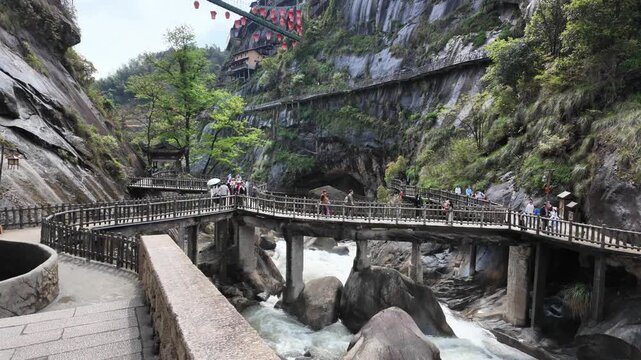 Wangxian Valley, China, 04 April 2026 ? Tourists cross a stone bridge over a rushing mountain river inside a dramatic rocky gorge with cliffside walkways and red lanterns in spring.