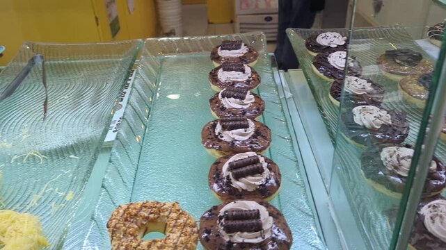 Close-up shot of various freshly baked chocolate and cream donuts displayed on glass trays inside a bakery showcase window.