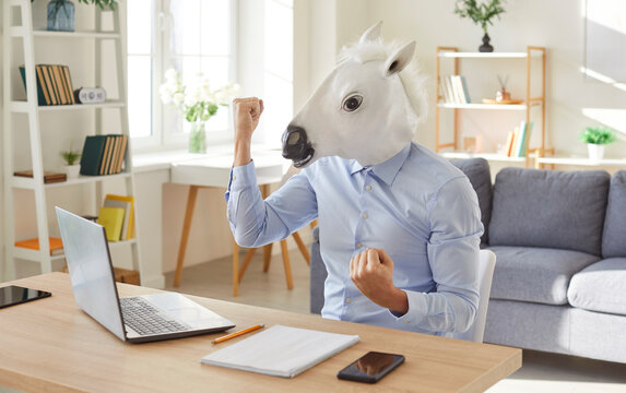 Excited man in formal shirt wearing white horse mask sitting at desk with laptop and celebrating success. Funny surreal office worker winning lottery, getting job offer and enjoying positive news.