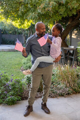 Mature African American man holding child girl in pink waving American flags on backyard patio