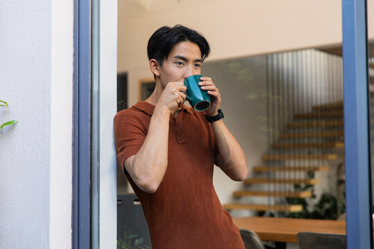 Asian man wearing rust polo, dark smartwatch sipping from teal mug in doorway, leaning, copy space