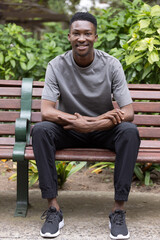 Obraz premium Adult African man sitting on wooden park bench wearing grey tee and black joggers, facing camera